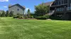Green lawn with houses, trees, shrubs, and a raised deck under a sunny blue sky with clouds.