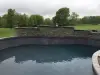 An empty backyard swimming pool with a stone wall, surrounded by grass and trees.