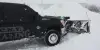 A black pickup truck with a snowplow clears deep snow in a snowy, winter setting.