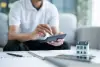 A person uses a calculator near documents and a small model house on a table.