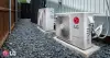Two LG outdoor air conditioning units sit on gravel beside a building and wooden fence.