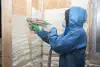 Person in protective gear spraying foam insulation onto wooden wall framing inside a building.