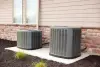 Two outdoor air conditioning units sit on a concrete pad next to a house with siding and brick.