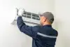A technician in uniform servicing a wall-mounted air conditioner unit indoors.