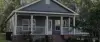 A small gray house with a front porch, white railings, and steps surrounded by plants.