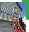 Person on ladders repairing damaged siding on a two-story house under a partly cloudy sky.