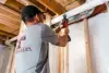 Person using a power drill on a pipe in an unfinished basement with exposed wooden beams.