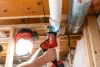 Person using a reciprocating saw to cut a white PVC pipe in a basement with exposed beams.