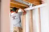 Man in a gray shirt installing white pipes in an unfinished basement ceiling.