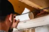Person installing a white PVC pipe fitting under a wooden ceiling with visible wiring nearby.