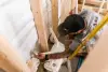 A worker installs white plumbing pipes in a framed wall of a building under construction.