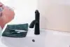 A black faucet on a white sink beside a black cloth and two markers, with hands at left.