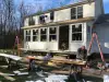 Two people work on the exterior of a white house under renovation, tools and materials scattered.