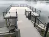 Wooden dock with black railings extending over calm water, seen from a ramp above.